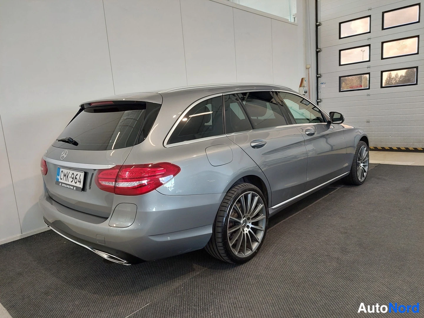 Takaviisto: Hopeanharmaa Mercedes-Benz farmariauto takaa kulmasta kuvattuna sisätiloissa, rekisterikilpi.