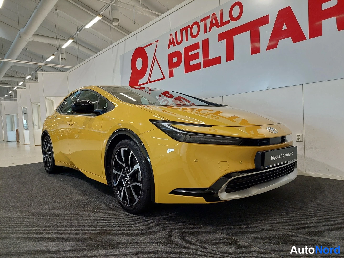 Etuviisto, nokka oikealle: Auton etukulma sisätiloissa; keltainen koripaneeli, konepelti, etugrilli ja Toyota Approved -kyltti näkyvät, tummat kevytmetallivanteet.