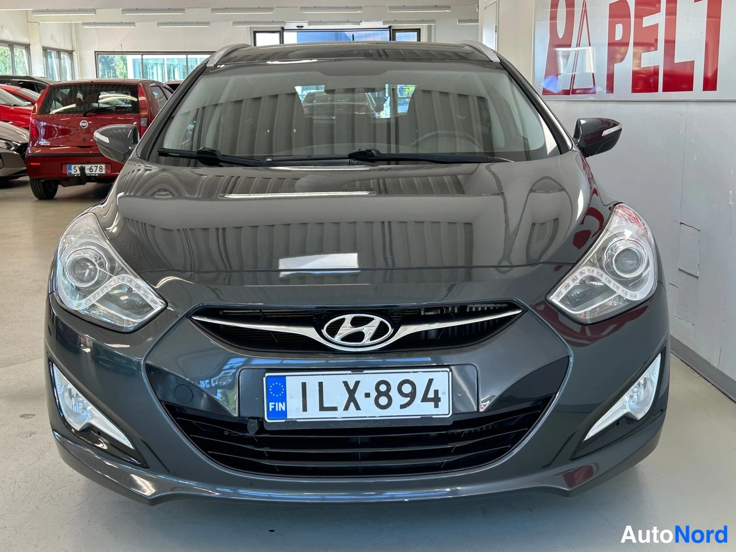 Edestä: Tummanharmaan farmariauton keula suoraan edestä, Hyundai-merkki ja rekisterikilpi näkyvät.