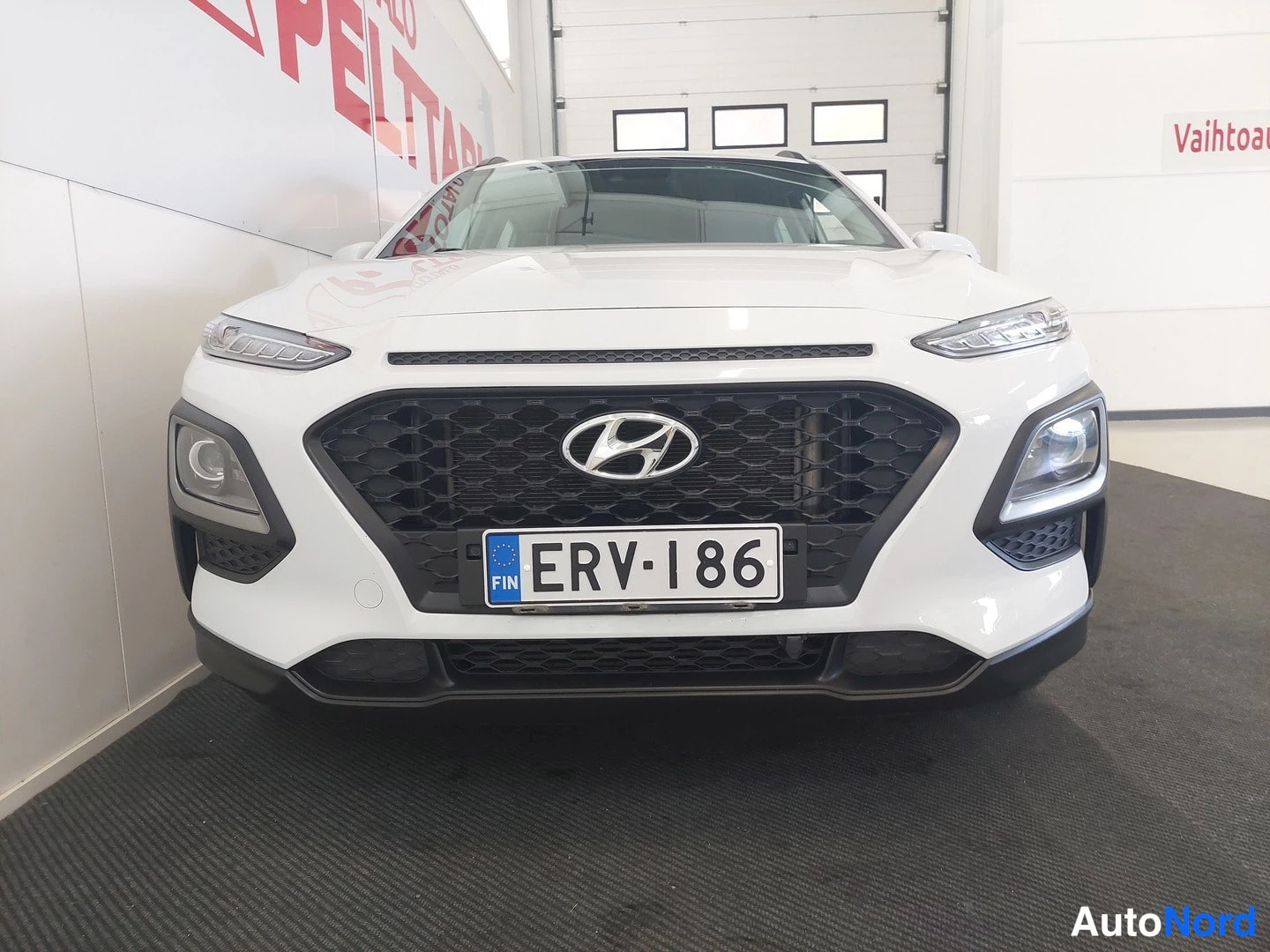 Edestä: Hyundai Kona -mallin etupää suoraan edestä valokuvattuna sisätiloissa; valkoinen kori, musta etusäleikkö ja rekisterikilpi näkyvät.