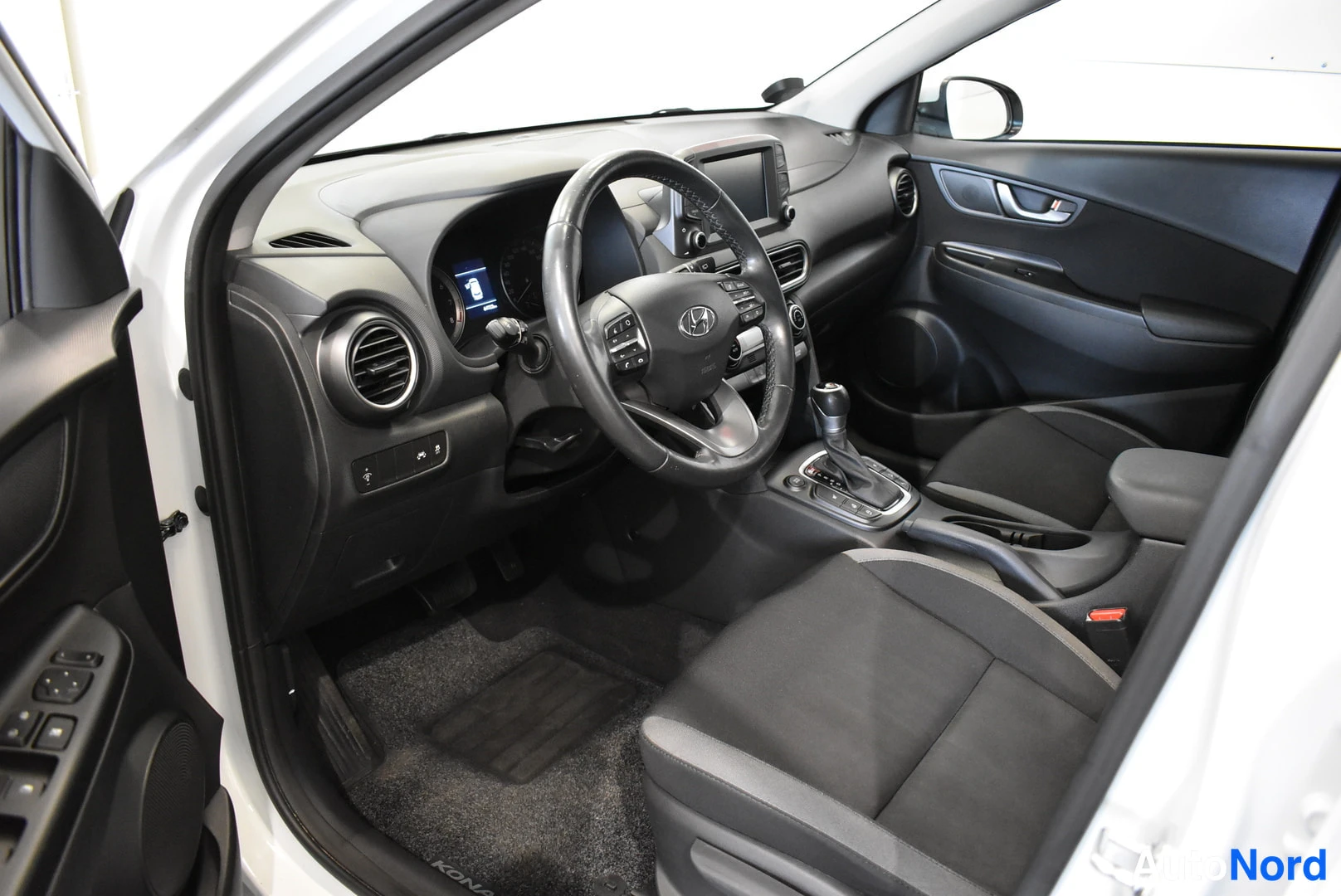 Muu: Hyundai Kona -mallin ohjaamo kuvattuna avatusta kuljettajan ovesta: musta verhoilu, ohjauspyörä, keskikonsoli ja etupenkit näkyvät.