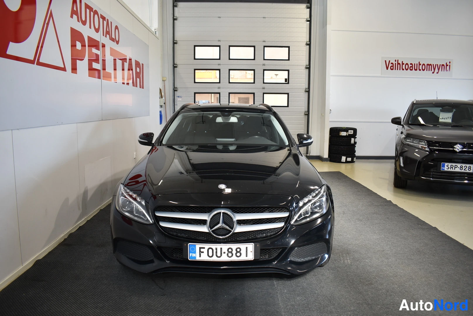 Edestä: Mustan Mercedes-Benzin etupää suoraan edestä; takaosa peilinä konepellissä, rekisterikilpi.