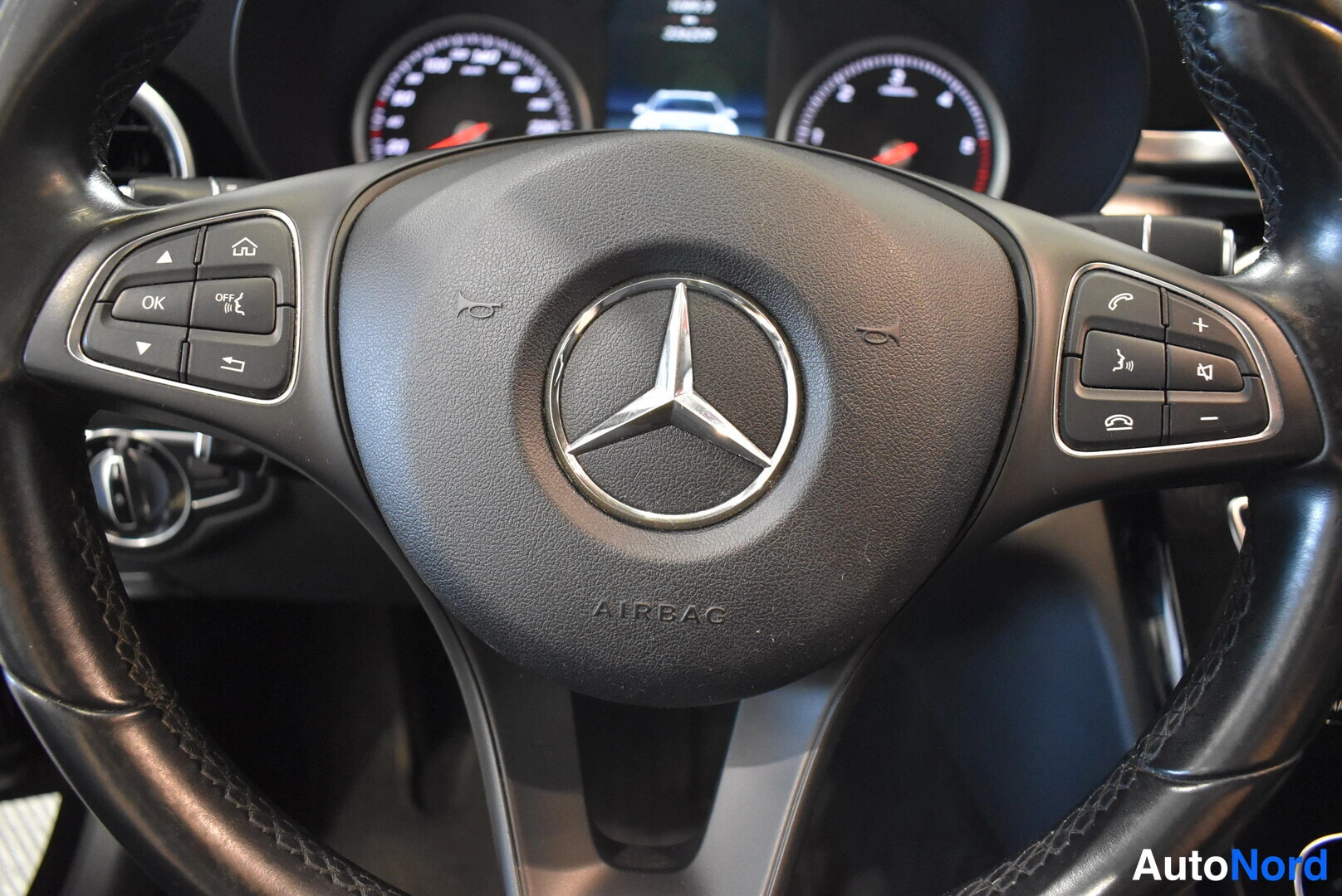 Muu: Mercedes-Benzin ohjauspyörän lähikuva: merkki keskellä ja näppäimet vasemmassa ja oikeassa puolessa, mittaristo taustalla.