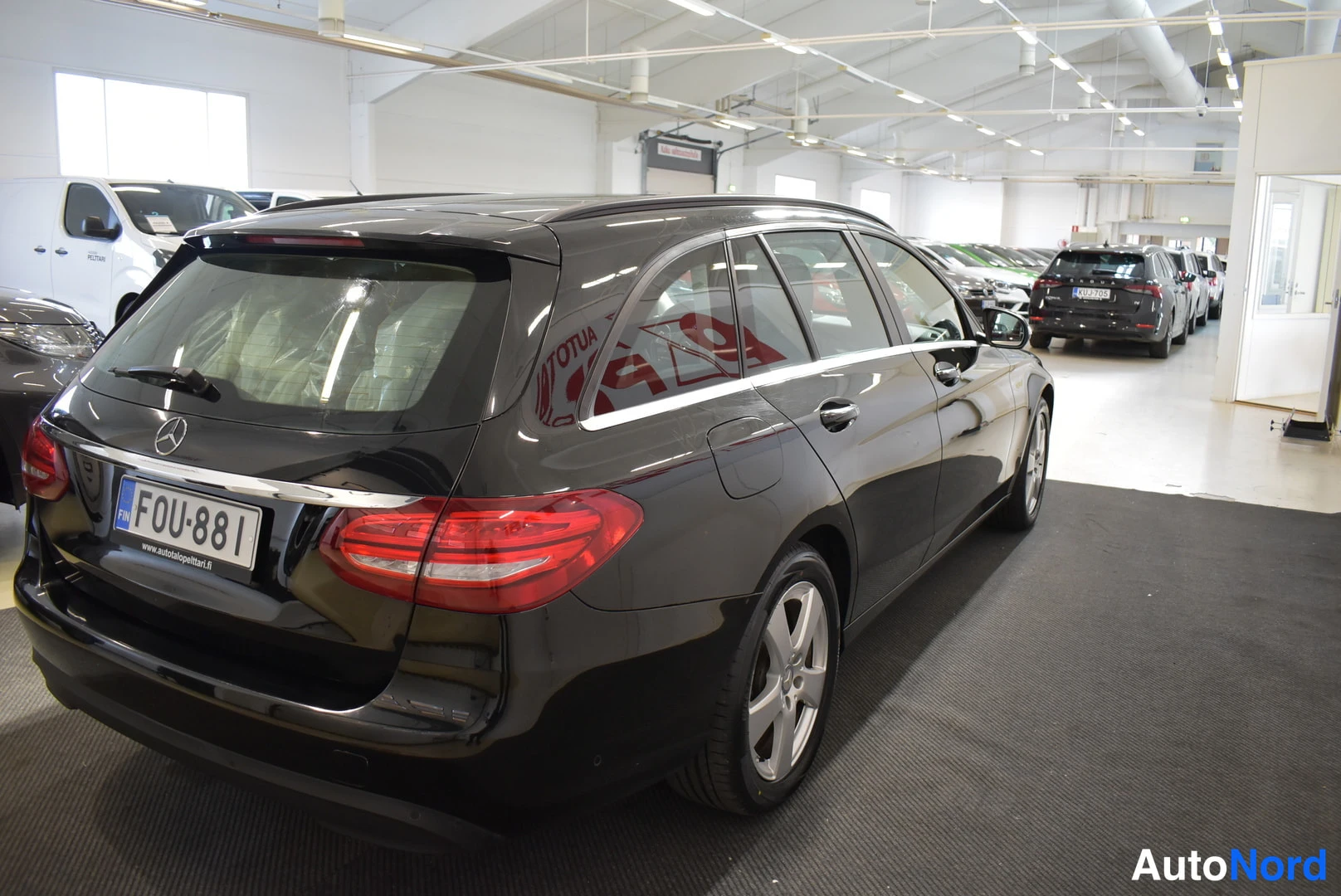 Takaviisto: Musta Mercedes-Benz -farmari vasemmalta takaa kuvattuna autosalissa; rekisterikilpi ja pölyä suojaava läpinäkyvyyttä takaluukun lasissa.