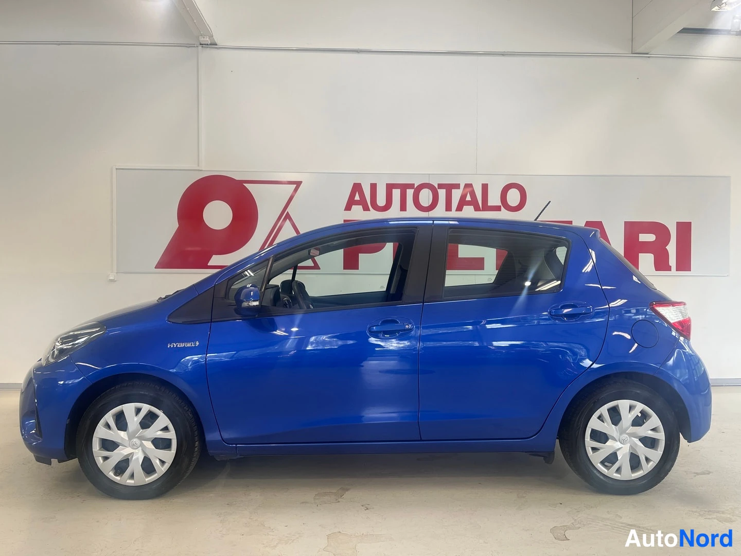Sivulta: Sivukuvasininen Toyota Yaris -henkilöauto oikealta sisätiloissa, neljä ovea ja HYBRID-kylkikyltti näkyvissä.