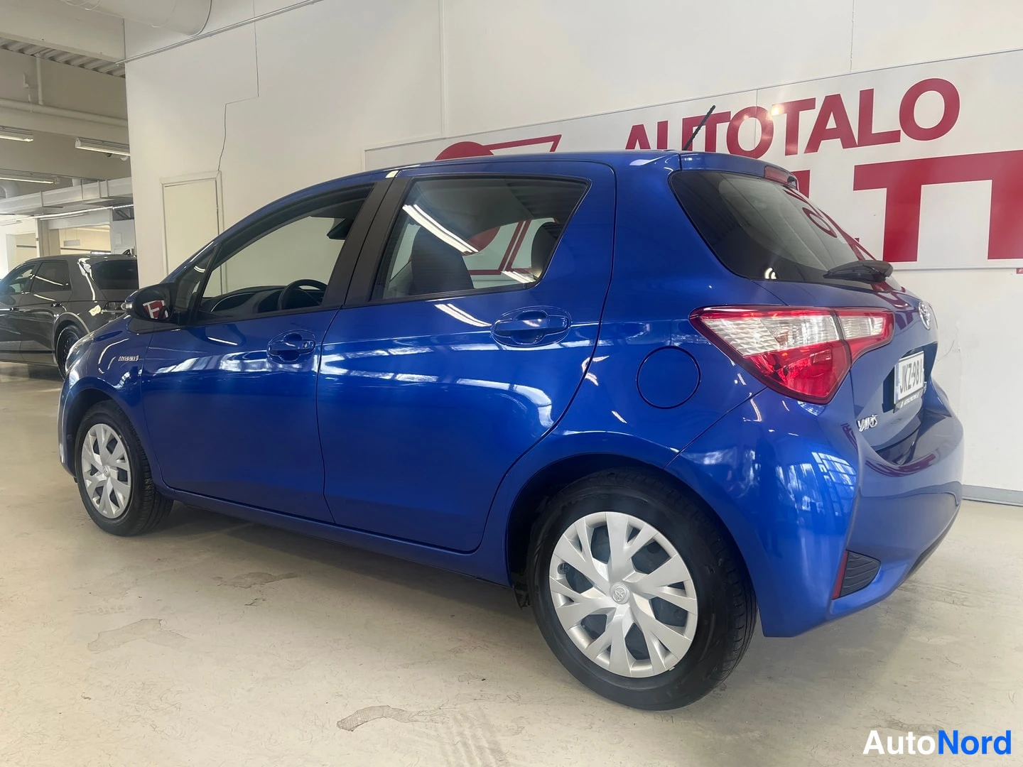 Takaviisto: Takimmainen vasemmanpuolinen 3/4-näkymä sininen Toyota Yaris sisätiloissa, takavalot ja rekisterikilpi näkyvissä.