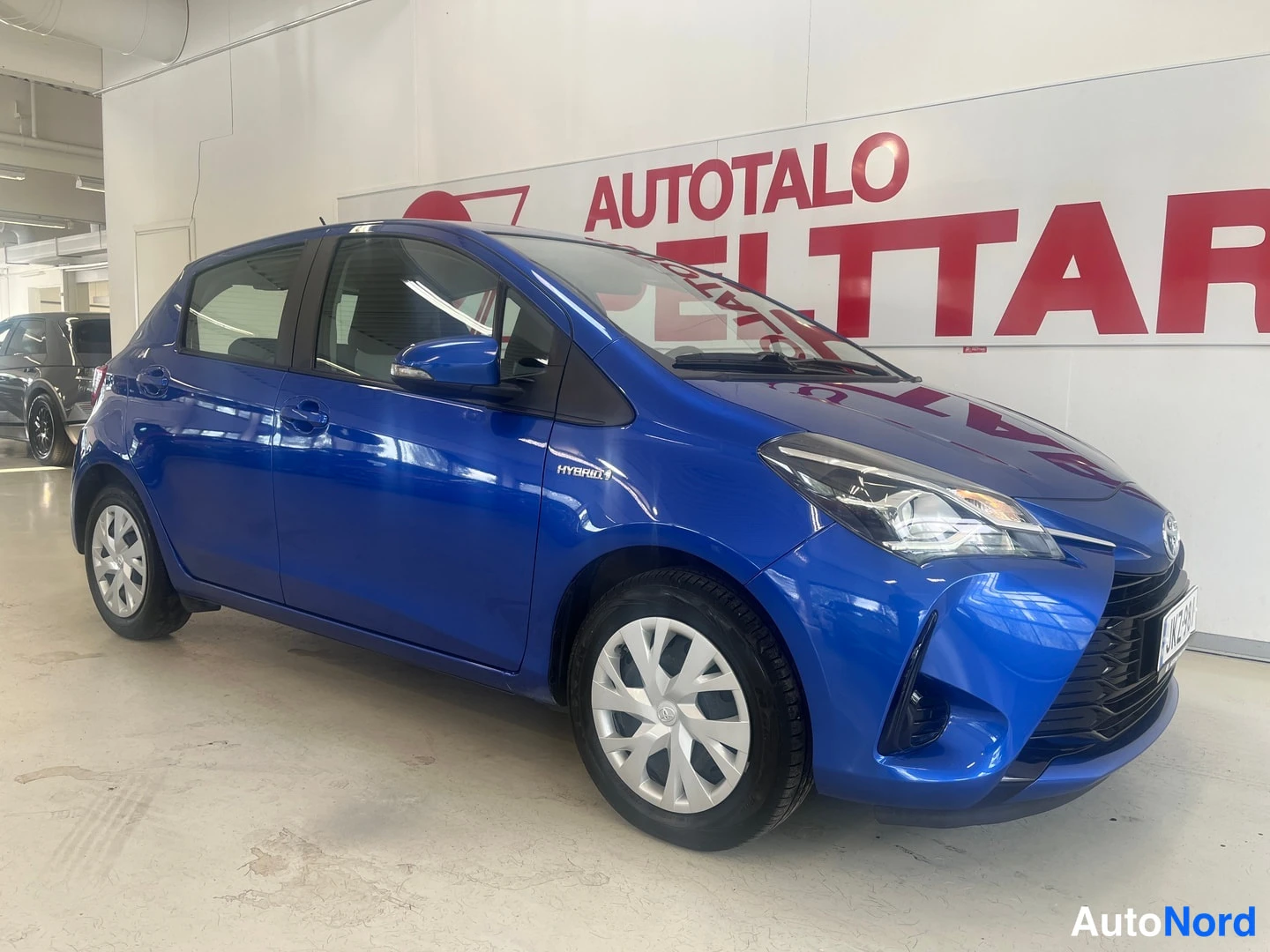 Etuviisto, nokka oikealle: Sinisen Toyota Yaris -henkilöauton etuosa kolmasosakulmasta keula oikealla, kuvattu sisätiloissa.