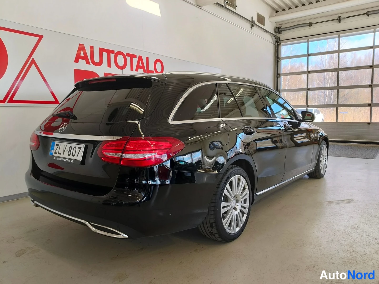 Takaviisto: Musta Mercedes-Benz C farmari takaa vinosti sisätiloissa, rekisterikilpi.