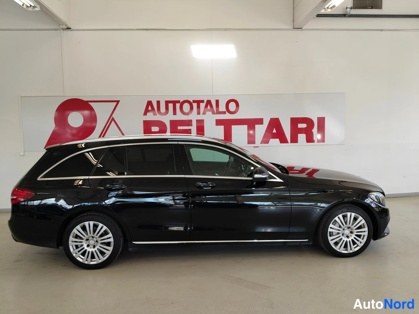 Etuviisto, nokka oikealle: Mustan Mercedes-Benz C-farmarin sivuprofiili hallissa, hopeiset vanteet ja kattokaaret.