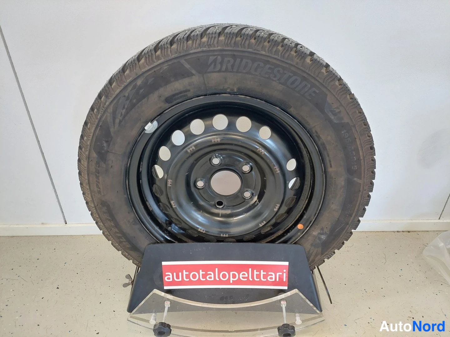 Yksityiskohta: Bridgestone-nastarenkaan päällinen mustalla teräsvanteella näyttöpöydällä, jonka kyljessä on punainen 'autotalopelttari'-merkki.