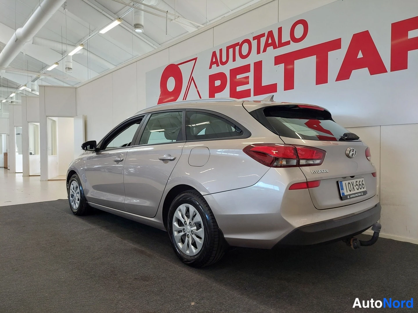 Takaviisto: Hyundai i30 Farming rear three-quarter view from the left in showroom, silver body, roof rails and tow hitch visible, license plate 10X-566.