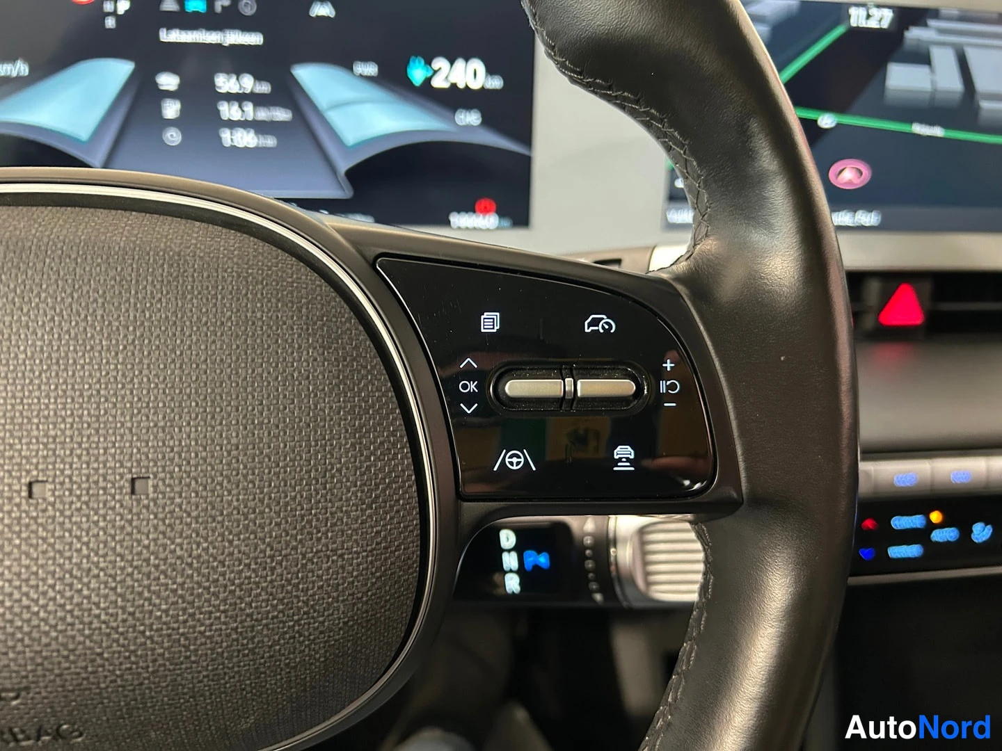 Muu: Hyundai IONIQ 5:n ratin oikean pöykärin äänenvoimakkuus- ja toimintopainikkeet lähikuvassa, taustalla instrumenttinäyttö ja infotainment-näyttö navigaatiokartal
