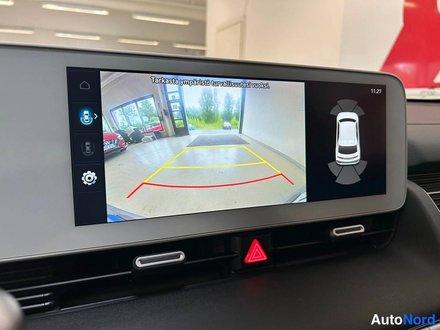 Muu: Hyundai IONIQ 5:n infotainment-näyttö kääntökuvanäkymässä tekstillä 'Tarkasta ympäristö turvallisuutesi vuoksi.', alla kojelautaan tuuletusaukot ja varoitusvalo