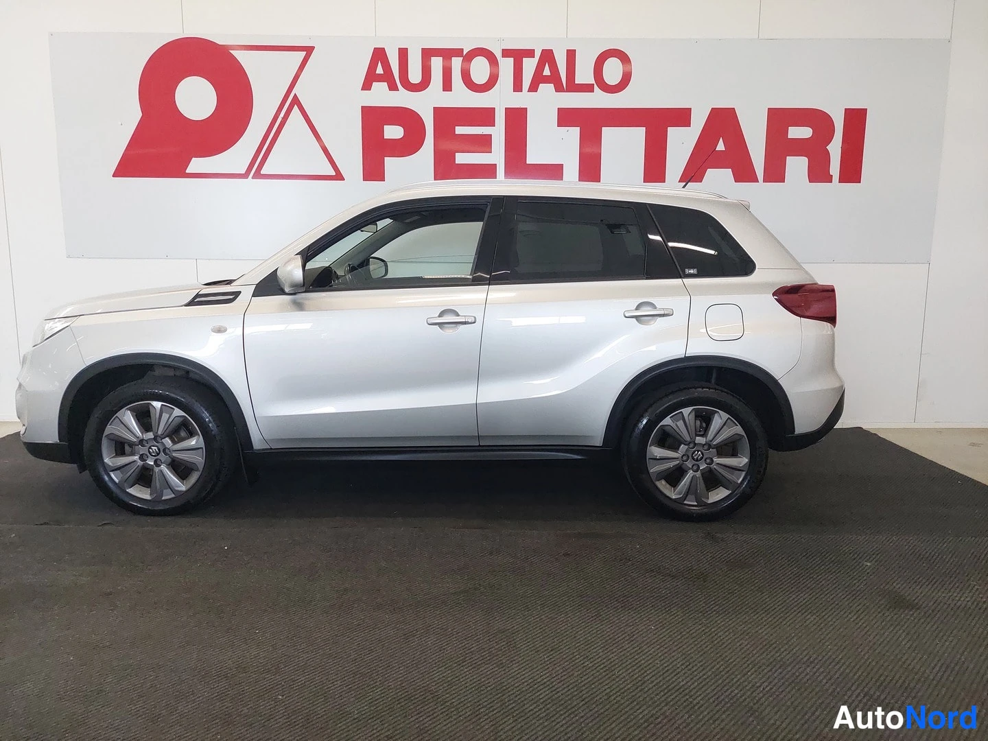 Takaviisto: Suzuki Vitara takaviisto valoisassa sisätilassa; hopeinen kori, takavalot ja takapuskurin musta muovi tunnus. Taustalla punavalkoinen 'AUTOTALO PELTTARI' -kyltt