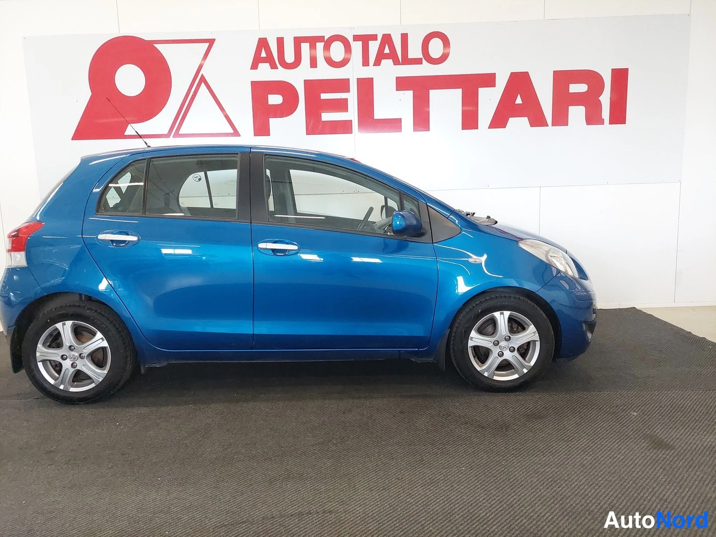 Etuviisto, nokka oikealle: Sininen Toyota Yaris viistosti edestä oikealta kuvattuna myyjän tiloissa.
