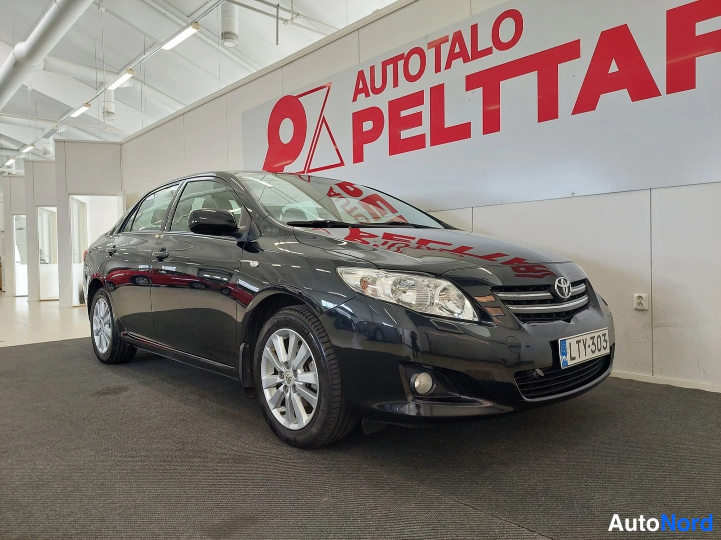 Etuviisto, nokka oikealle: Mustan Toyota Corolla -sedanin etuosa ja vasen kylki: etusäleikkö ja korostus Toyota-merkillä, rekisteritunnus, sumuvalot ja valkoinen taustakangas AutoT
