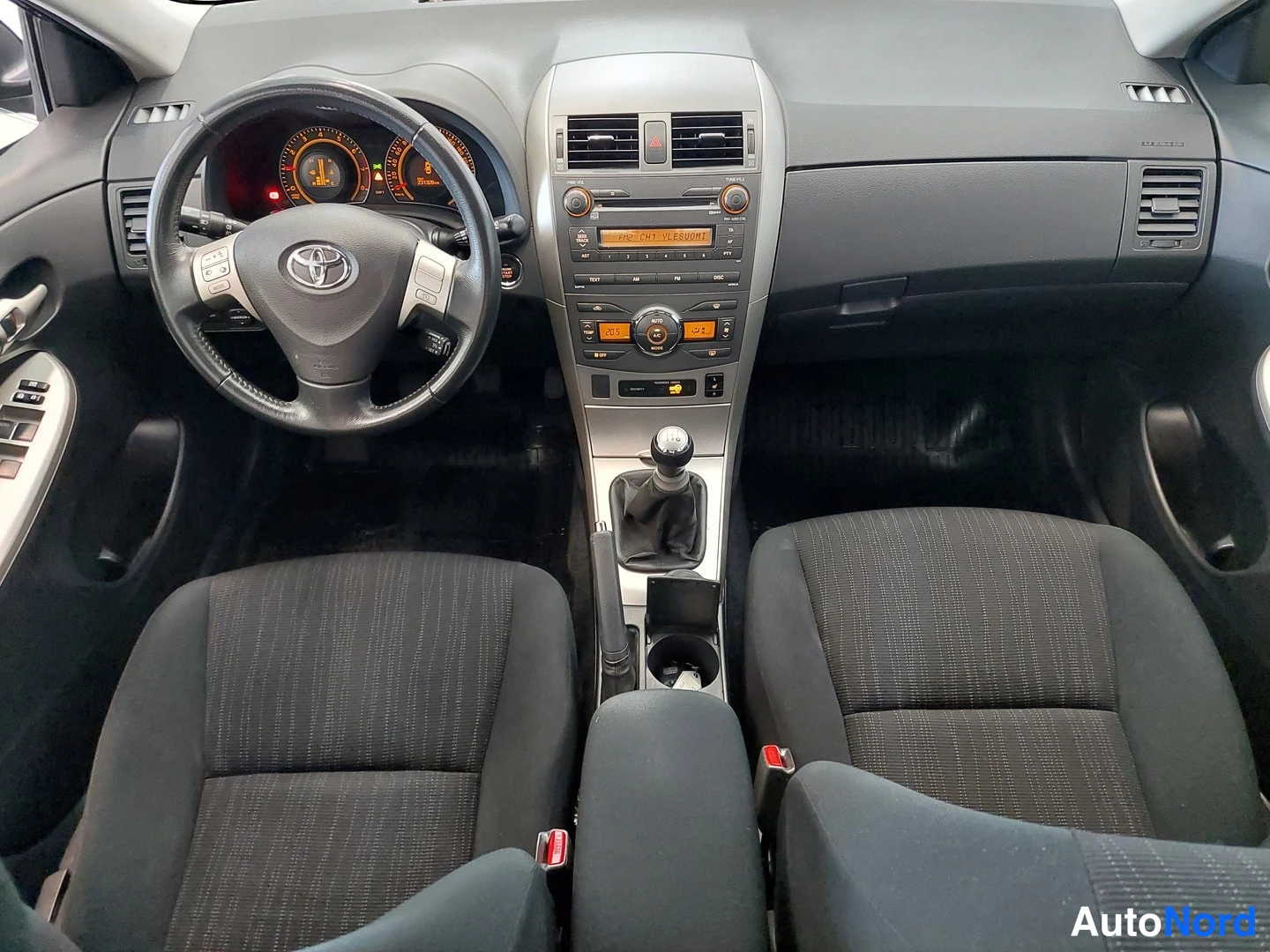 Muu: Ohjaamo eteenpäin: Toyota-ohjauspyörä, mittaristo, manuaalivaihteisto ja kaksi tummaa penkkiä.