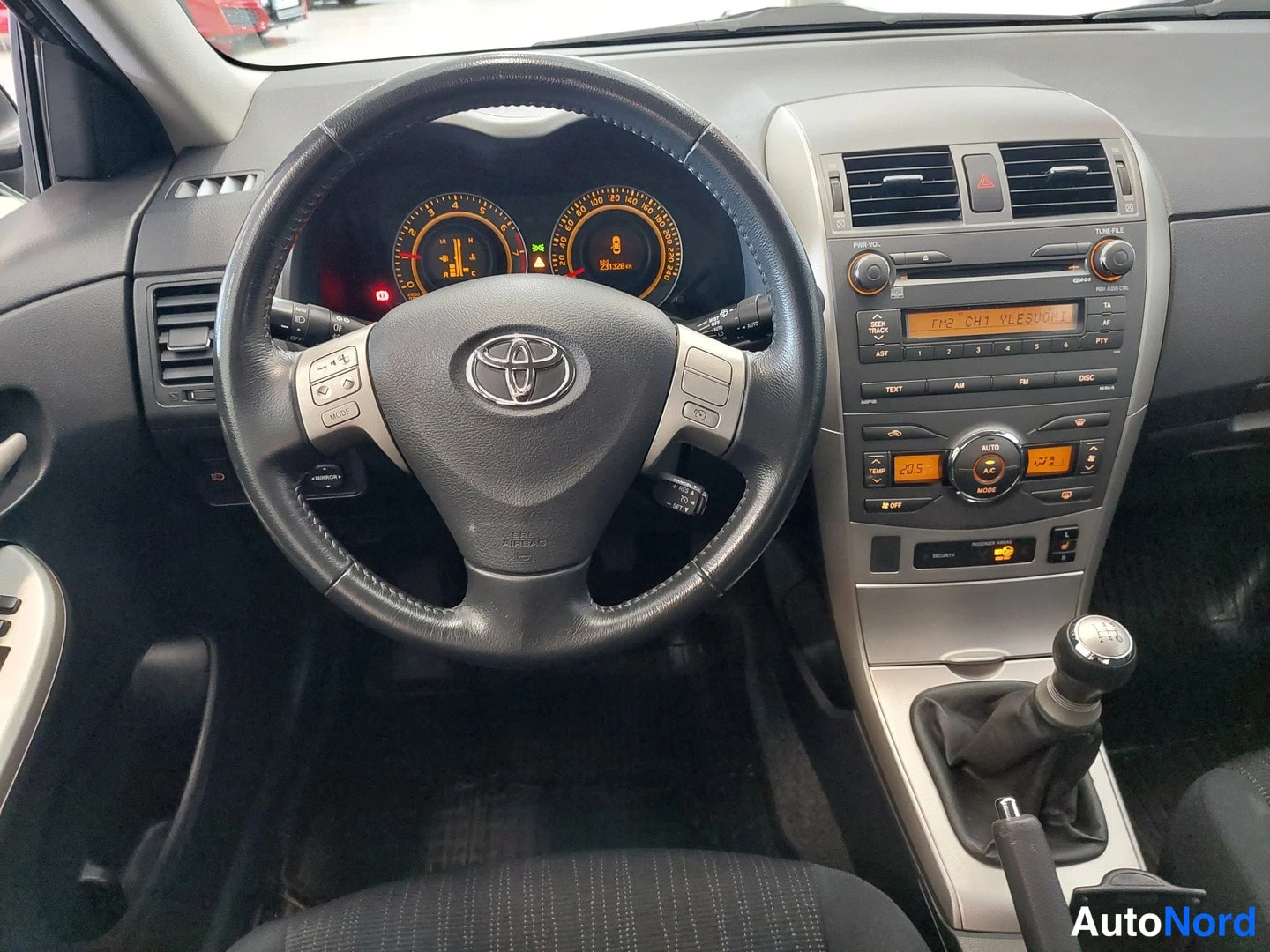 Muu: Ohjaamon tarkempi näkymä: Toyota-ohjauspyörä, mittaristo, radio ja automaattinen ilmastointipaneeli, manuaalivaihtokeppi.