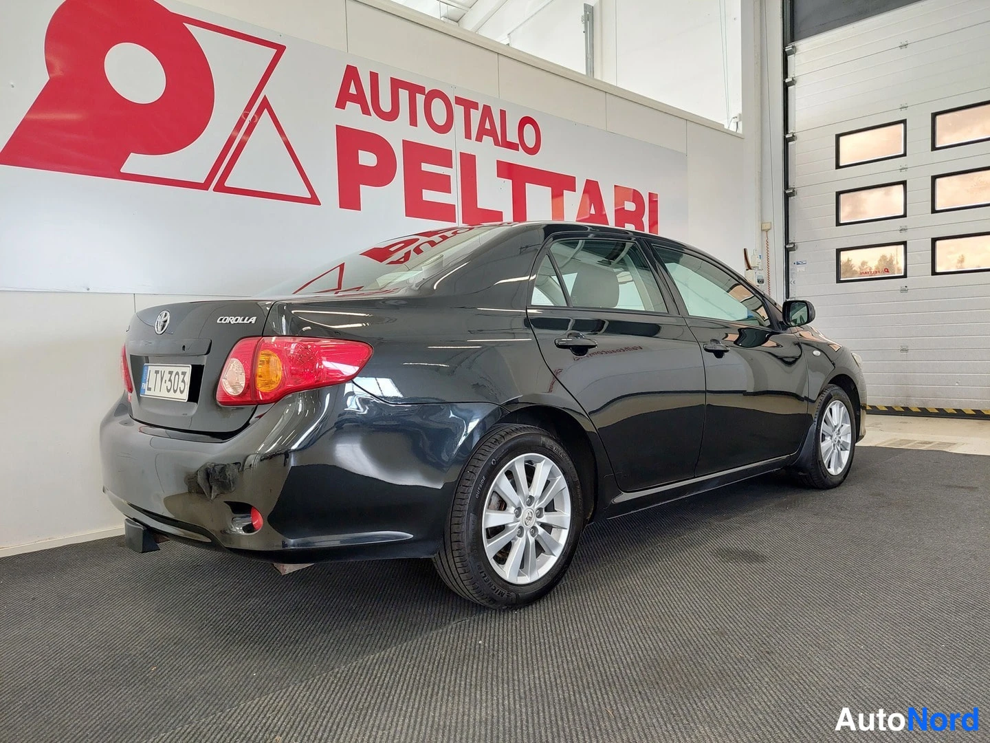 Takaviisto: Mustan Toyota Corolla -sedanin takaosa ja vasen kylki näkyvällä; taka- ja etupuskuri, rekisteritunnus ja paljon valokuvattu katsastushuoneen seinä AutoTalo Pelt