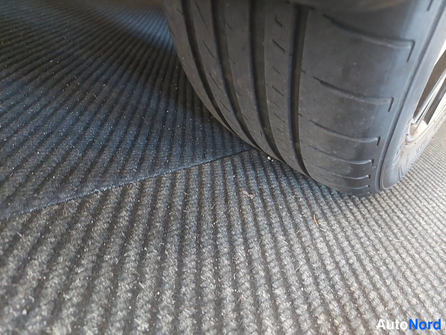 Yksityiskohta: Lähikuva auton renkaasta ja metallivanteen reunasta mattopohjaisella lattialla.