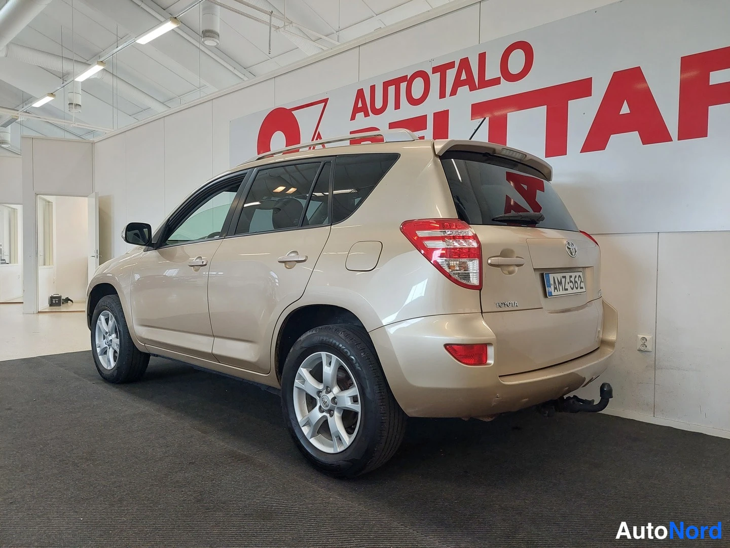 Takaviisto: Beige Toyota RAV4 takalokavälistä kuvattuna myyntihallissa, takaosan valot ja kylki näkyvissä, rekisterikilpi.
