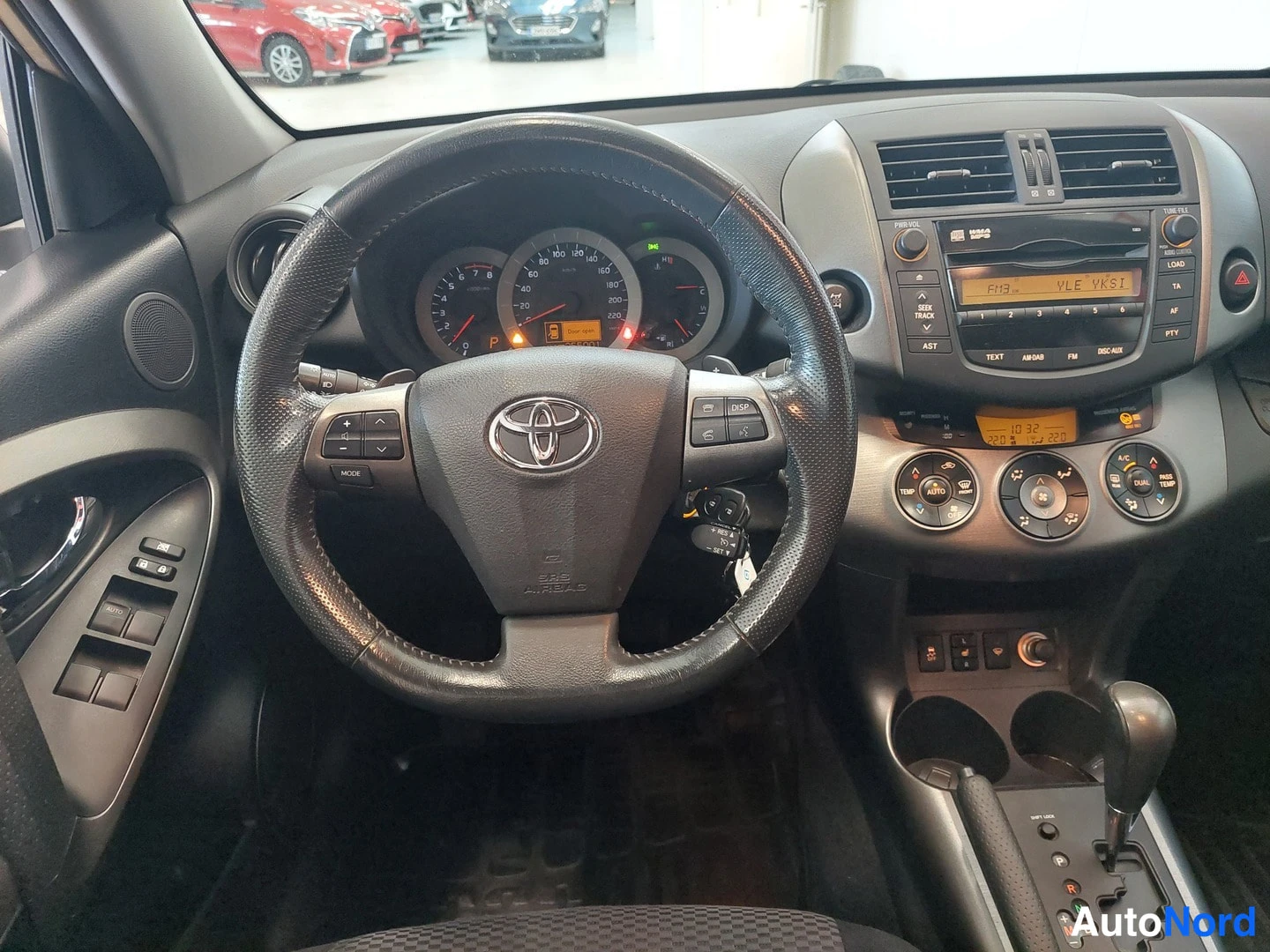 Muu: Toyota RAV4:n kojetaulu ja ohjauspyörä lähikuvassa, Toyota-merkki ja mittaristo näkyvät.