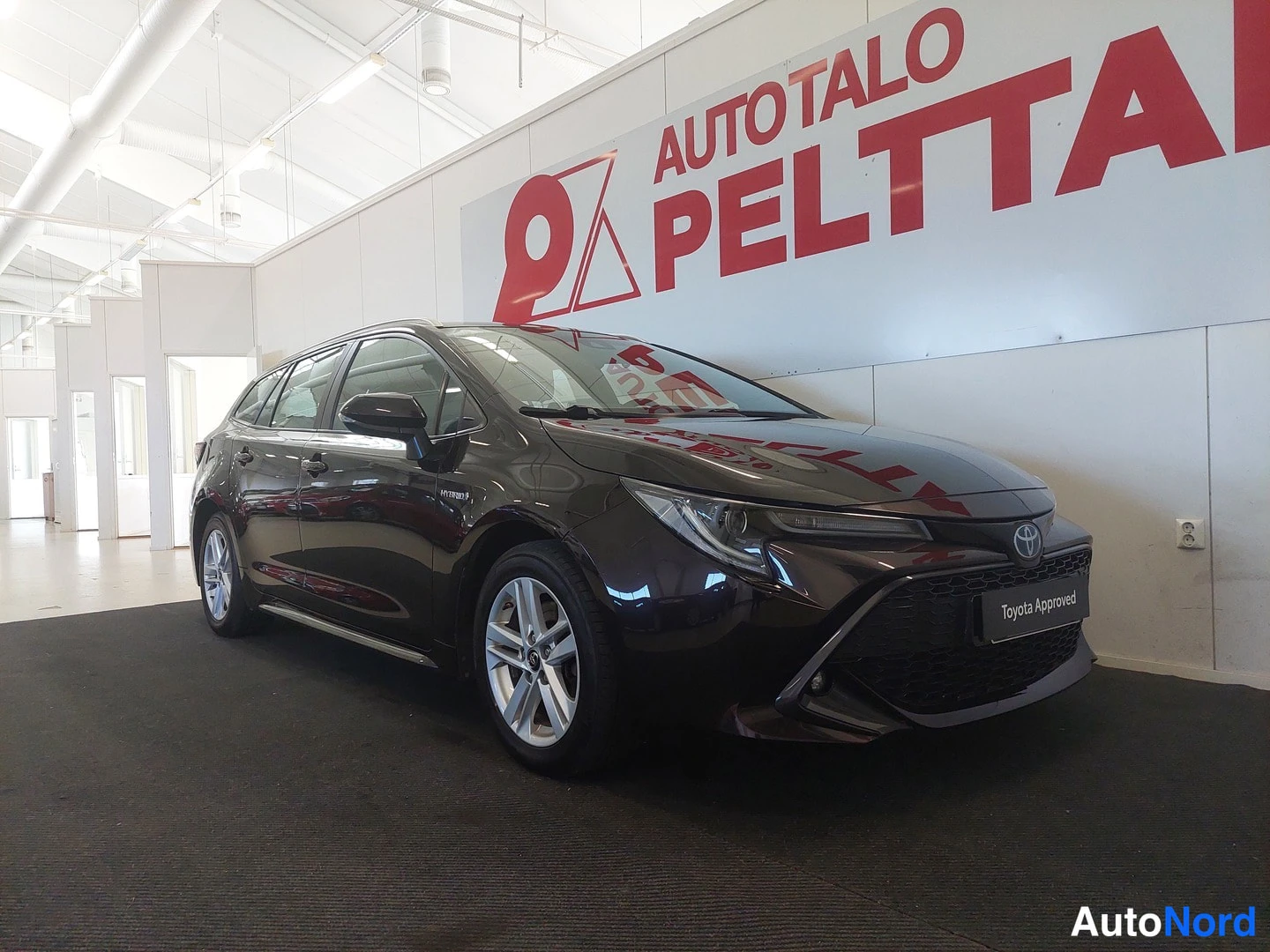 Etuviisto, nokka oikealle: Auto etu-37-näkymästä sisätiloissa; tummanruskea Toyota-farmari, Toyota-merkki keulalla ja Toyota Approved -kyltti etupuskurissa.