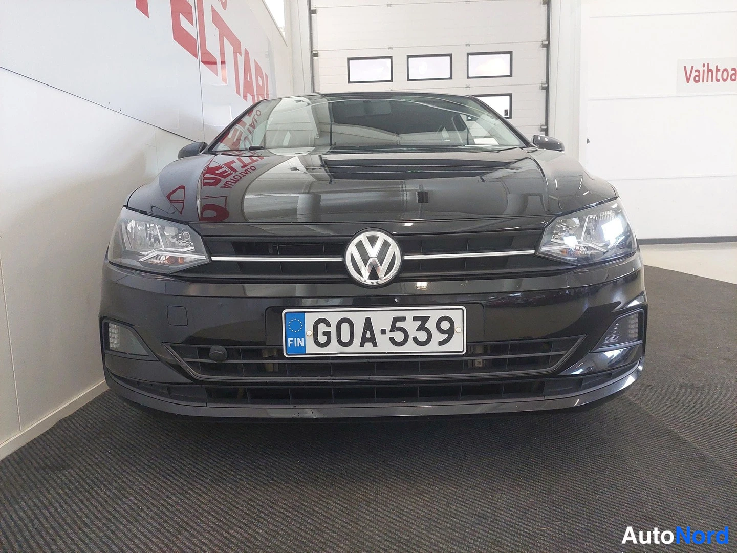 Edestä: Mustan henkilöauton etunäkymä; etupanssari, VW-merkki ja rekisterikilpi näkyvät sisätiloissa.