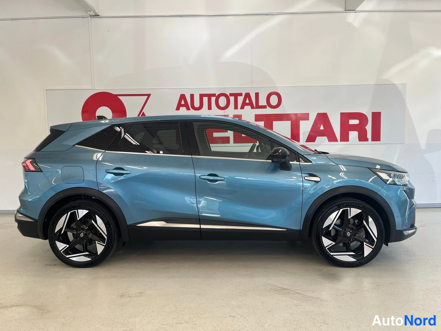 Etuviisto, nokka oikealle: Sinertävänharmaa Renault Symbioz sivulta oikealta nähtynä hallissa; keula ja etuvalo näkyvät hieman oikealla.