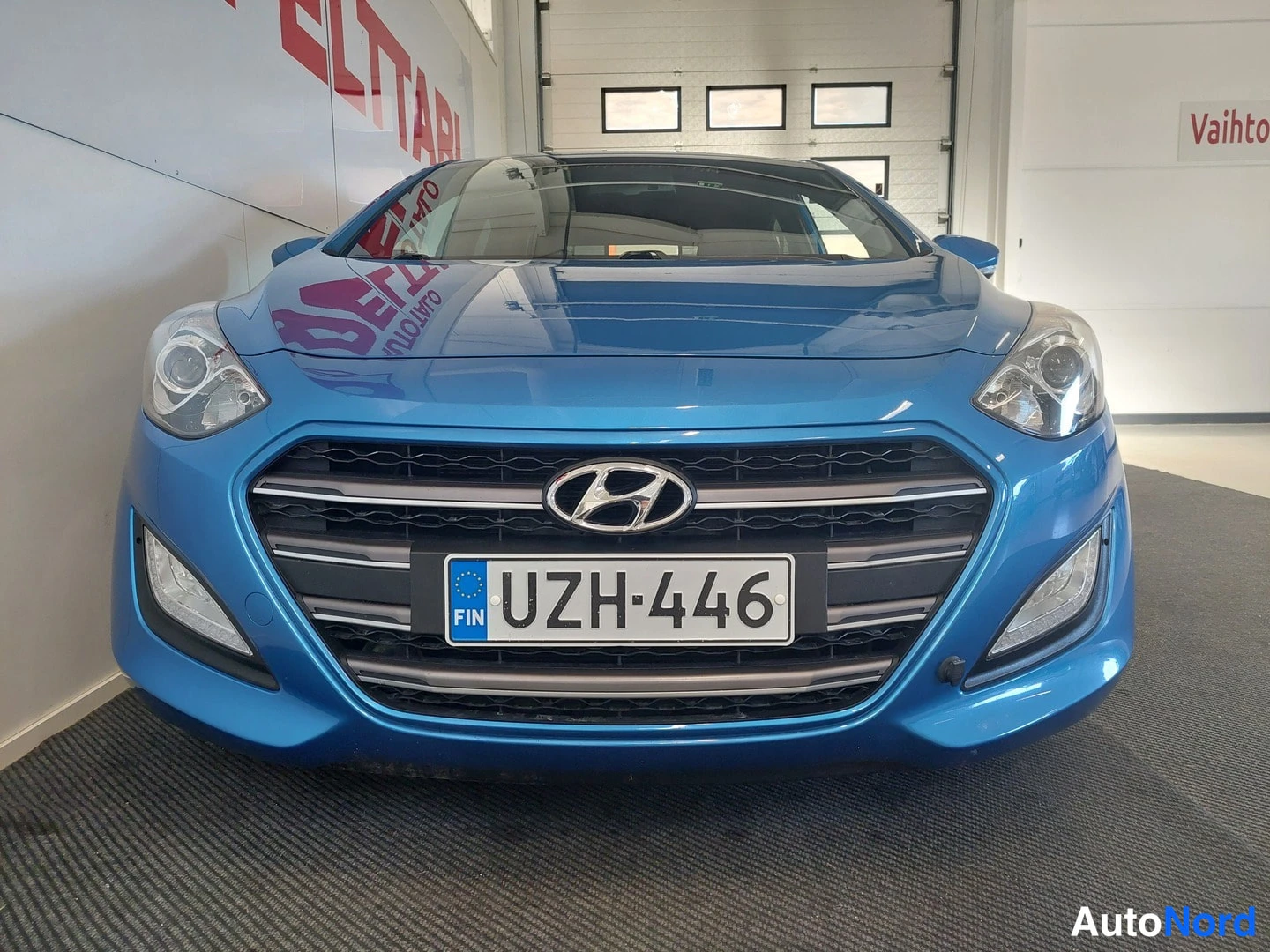 Edestä: Sininen Hyundai i30 edestäpäin kuvattuna sisätiloissa; etusäleikössä Hyundai-merkki ja rekisterikilpi.