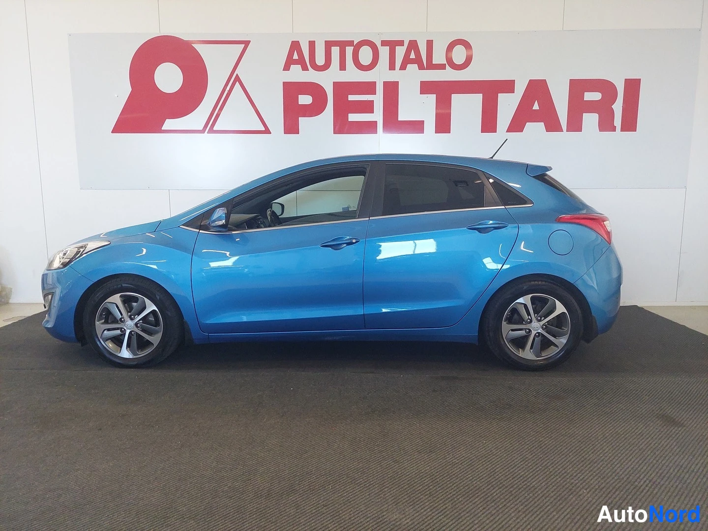 Etuviisto, nokka vasemmalle: Sininen Hyundai i30 -henkilöauto sivulta etuosa vasemmalle, sisätiloissa valkoisella taustalla.