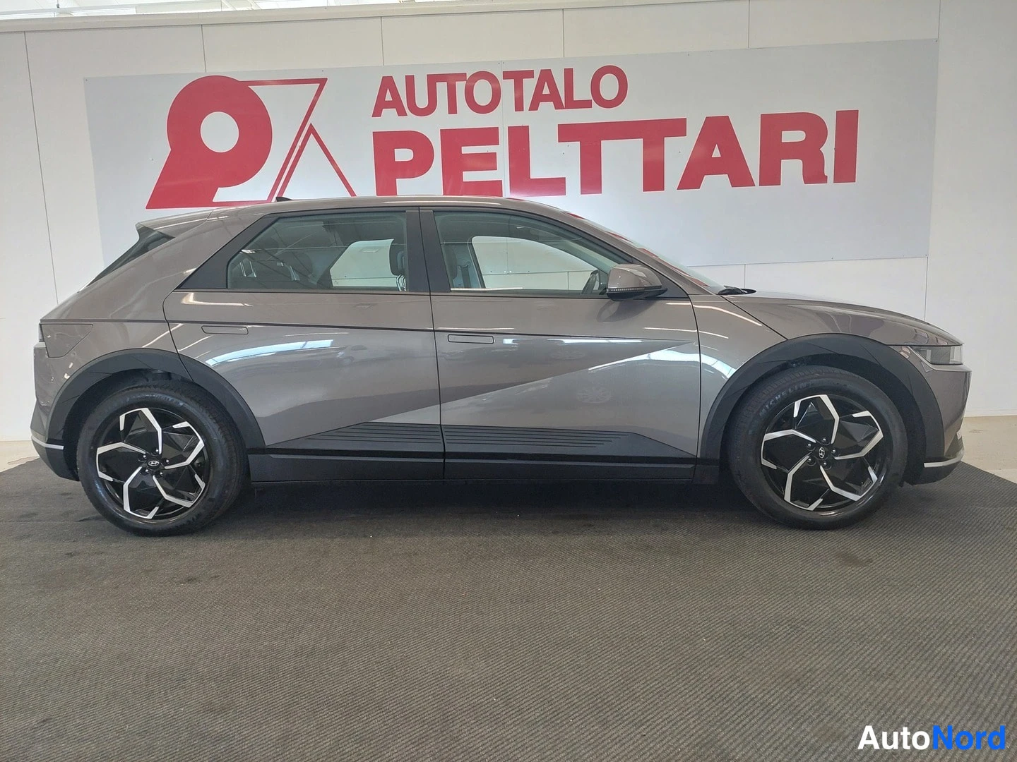 Sivulta: Hyundai IONIQ 5:n kylki suoraan sivulta, tummanharmaa kori ja muotoillut kevytmetallivanteet; taustalla AUTOTALO PELTTARI -mainoskyltti.