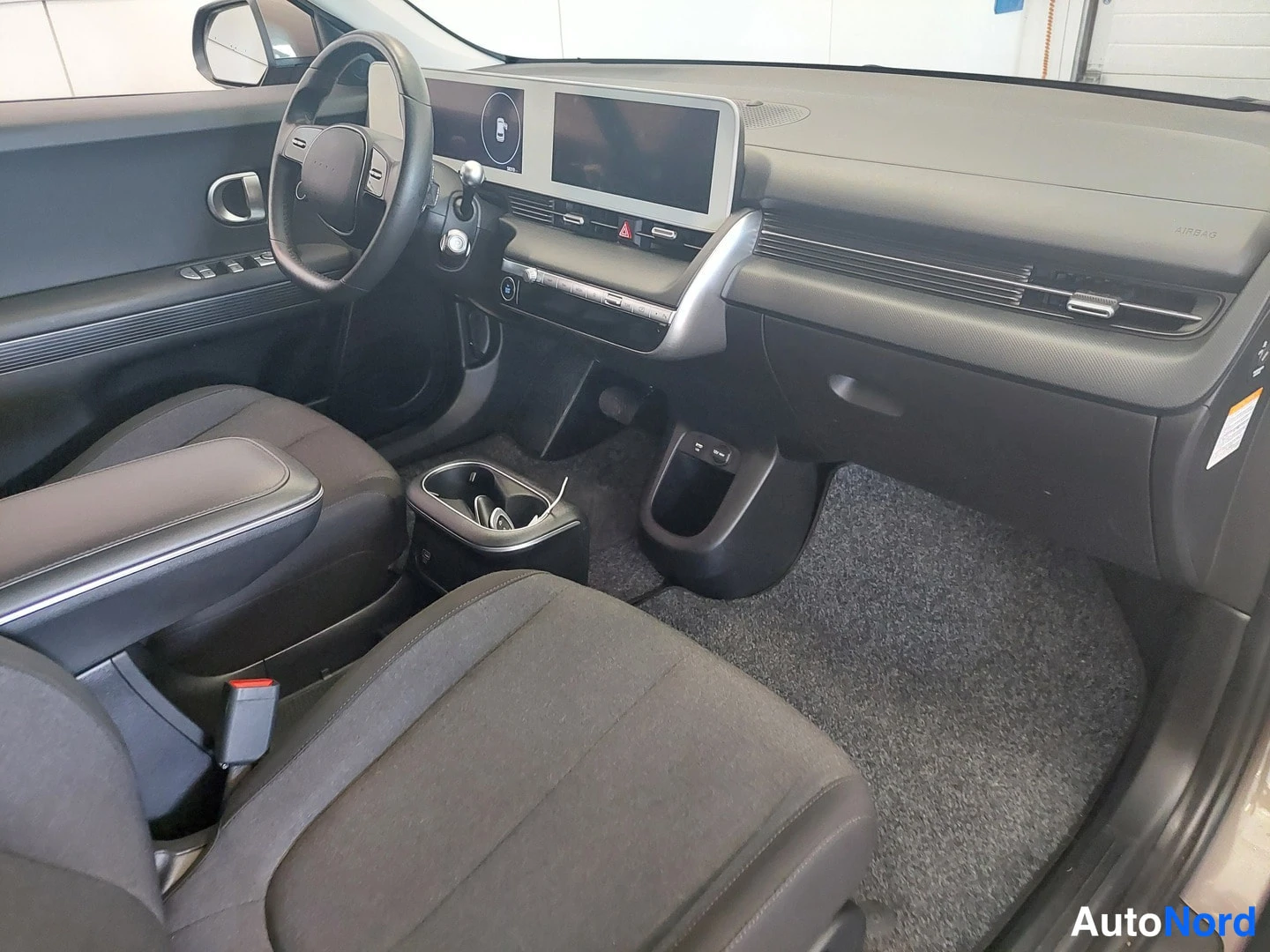 Muu: Hyundai IONIQ 5:n etuistuinpeili ja ohjaamon näkymä: tumma kangasverhoillut etuistuimet, keskikäytävä kosketusnäytöllä ja ilmastointipaneeleilla.