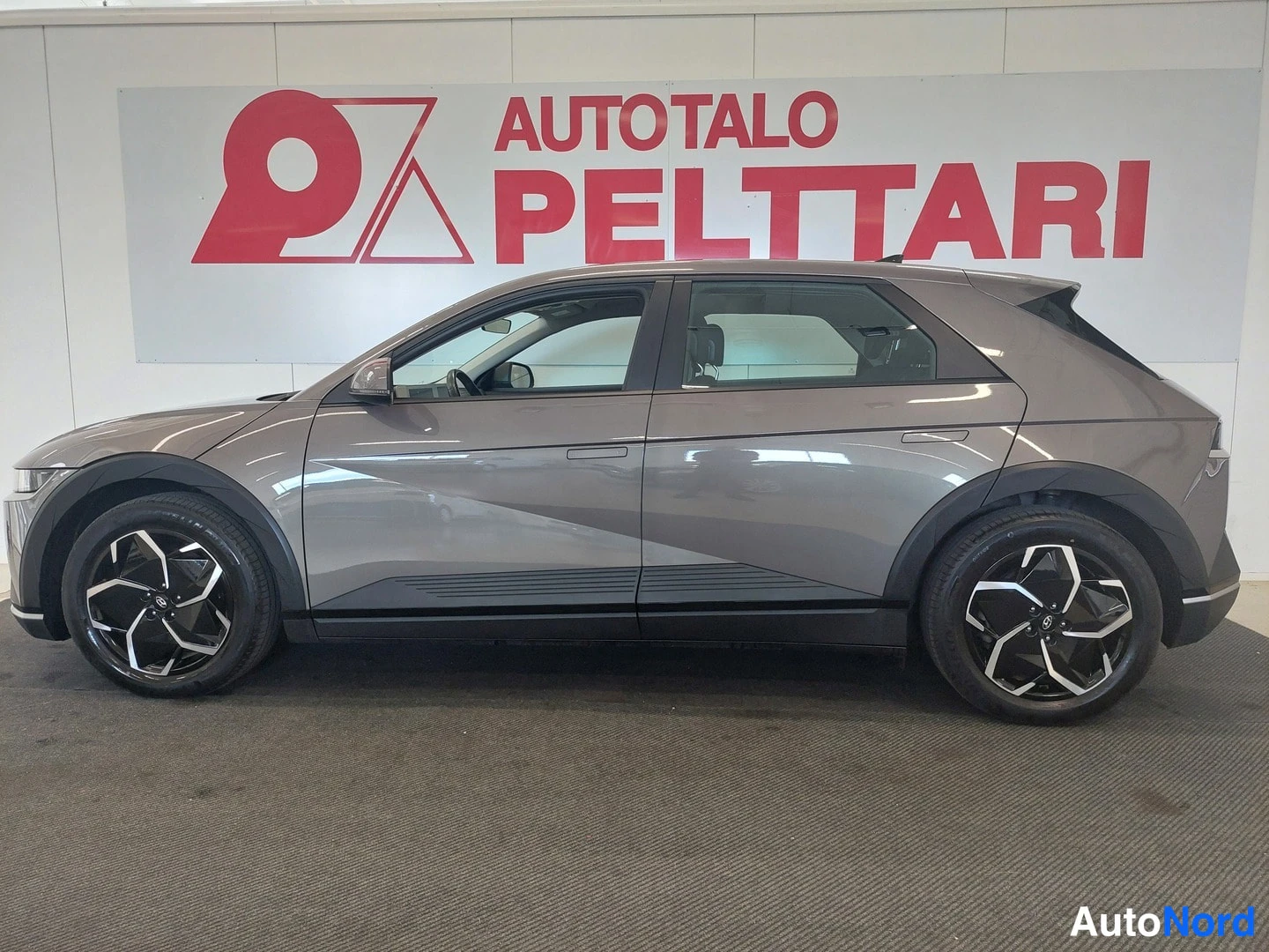 Etuviisto, nokka vasemmalle: Hyundai IONIQ 5 etukulmasta vasemmalta, tummanharmaa crossover-automaalilla sisätiloissa.