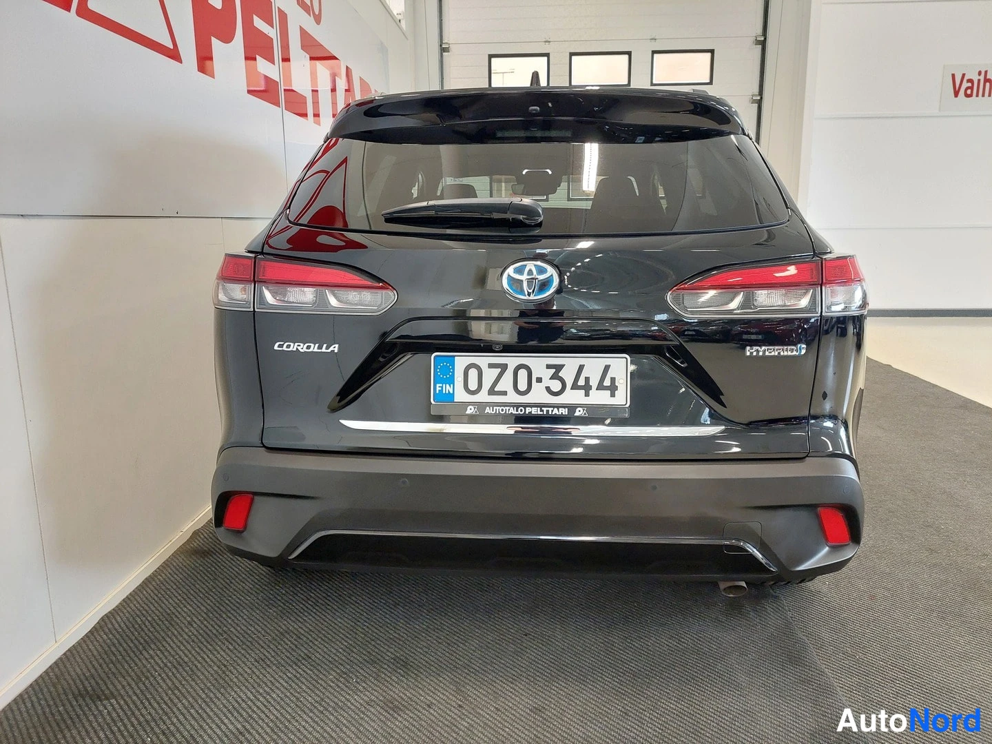 Takaa: Mustan Toyota Corolla Crossin takanäkymä sisätiloissa, rekisterikilpi ja HYBRID-merkintä takaluukussa.