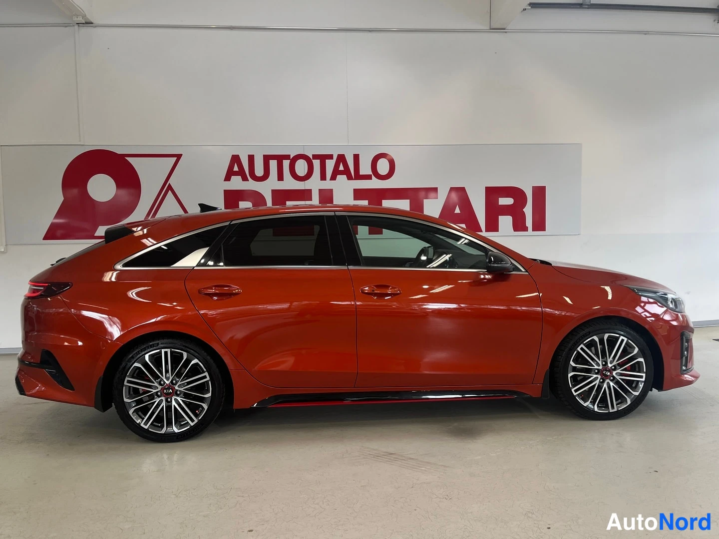 Sivulta: Punaisenoranssi Kia ProCeed sivusta kuvattuna: näkyvät molemmat ovet, mustat ikkunakehykset, alusta-paketit ja monisakeiset kevytmetallivanteet.