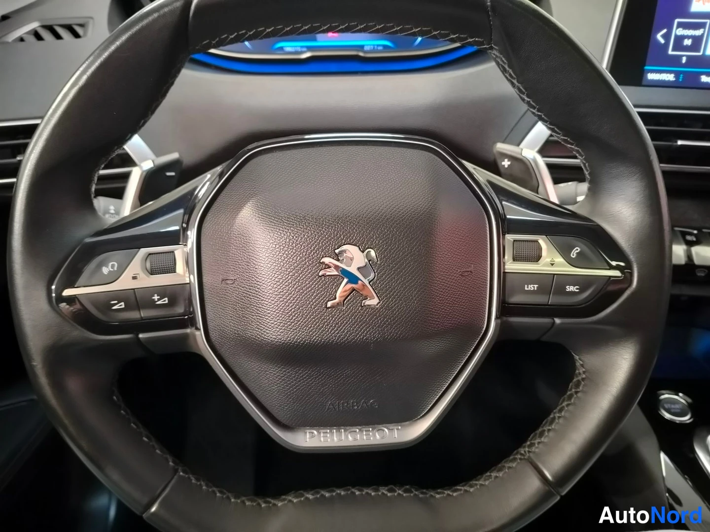 Muu: Musta Peugeot-ohjauspyörä leijonalogolla, AIRBAG-tekstillä ja monitoimipainikkeilla; mittaristö ja osa keskiconsolessa näkyvät taustalla.
