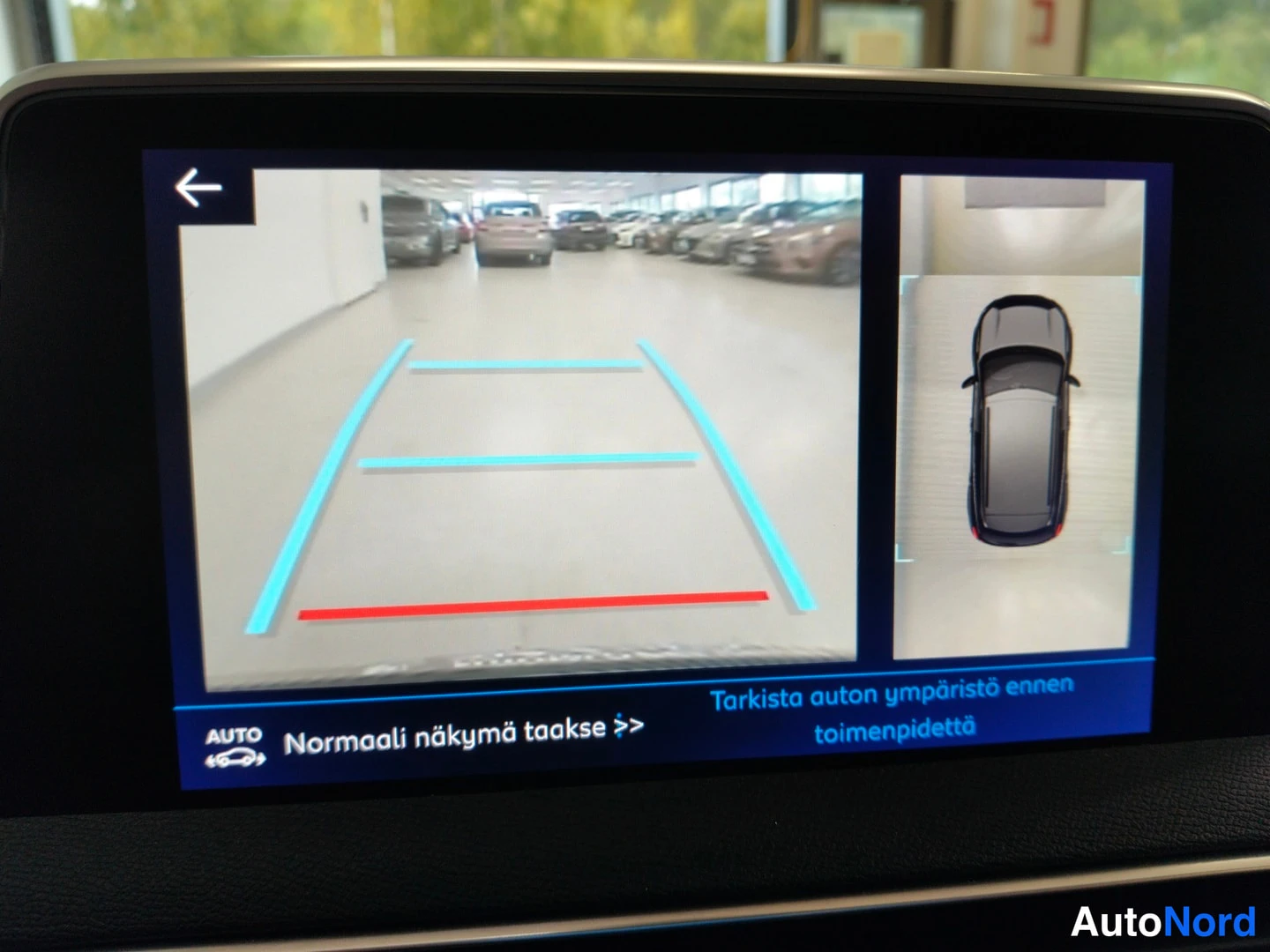 Yksityiskohta: Infotainment-näyttö näyttää peruutuskameran kuvan ohjausviivoilla ja tekstin 'Normaali näkymä taakse' sekä varoituksen 'Tarkista auton ympäristö ennen toimenpid