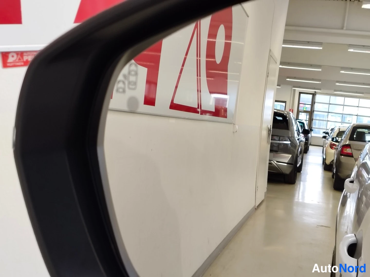 Muu: Ulkopeilin takaa kuvattu näkymä autosalon käytävään, jossa rivissä autoja; vasemmalla valkoinen seinä punaisin mainoskyltein.