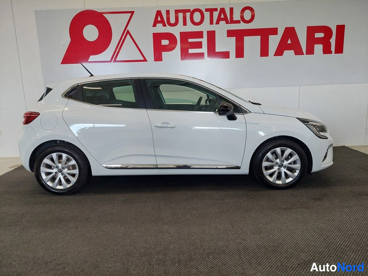 Sivulta: Valkoinen Renault Clio -viistoperä sivulta kuvattuna autosalon lattialla, Autotalo Pelttari -kyltti taustalla.