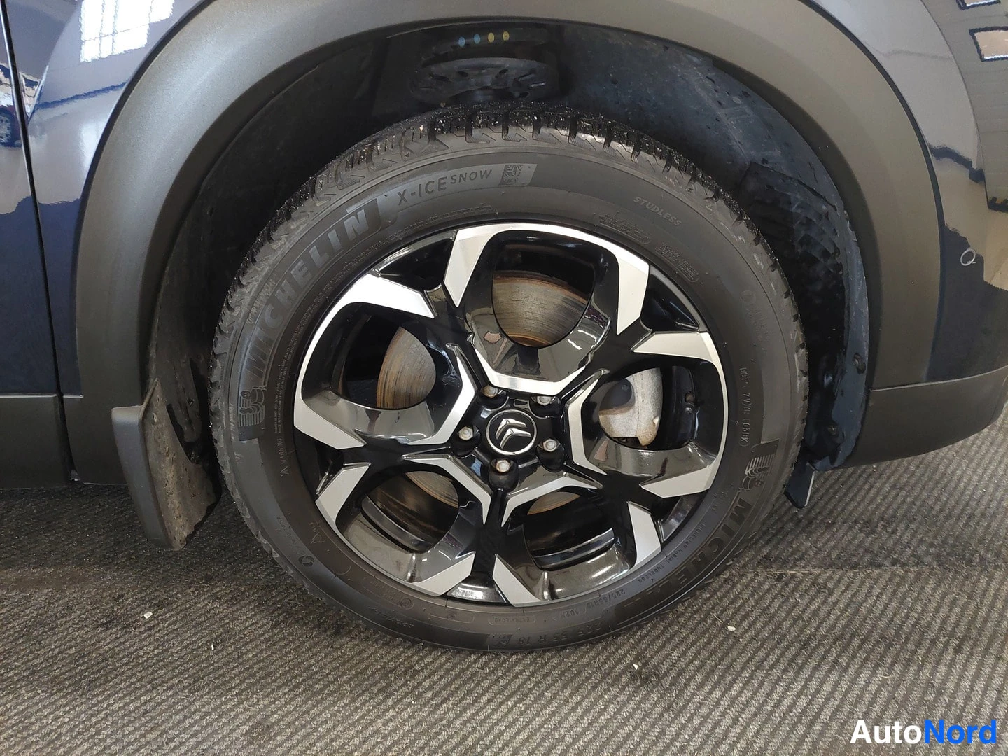 Yksityiskohta: Lähikuva mustasta kevytmetallivanteesta ja Michelin X-ICE SNOW -renkaasta tumman auton pyöräkaarella sisätiloissa.