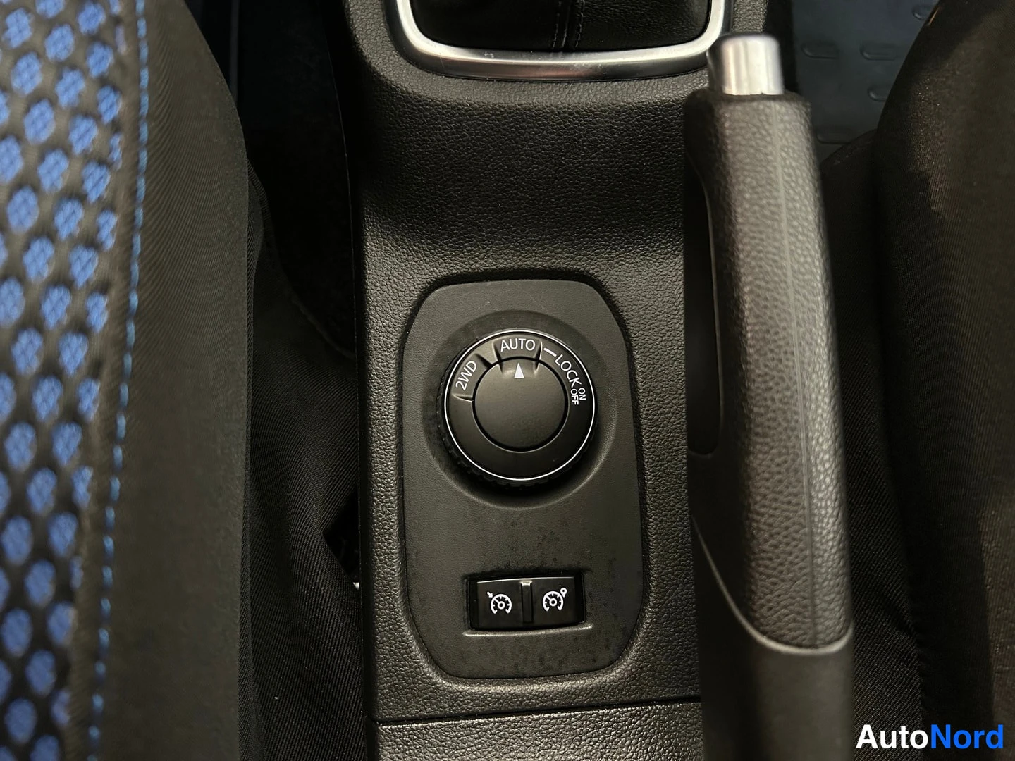 Muu: Sisätilakuva: kojelaudan kytkinnuppi, jossa tekstit 2WD, AUTO ja LOCK, sekä kaksi pientä kytkintä ja käsijarruvipu.