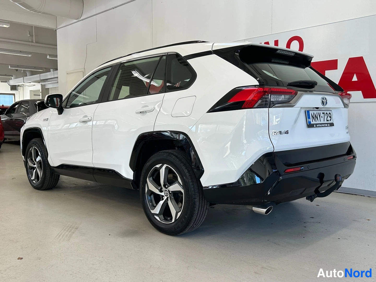 Takaviisto: Valkoisen Toyota RAV4:n takaosan kolmannesvasen näkymä sisätiloissa; rekisterikilpi, mustat puskurikulmat ja vetokoukku näkyvissä.