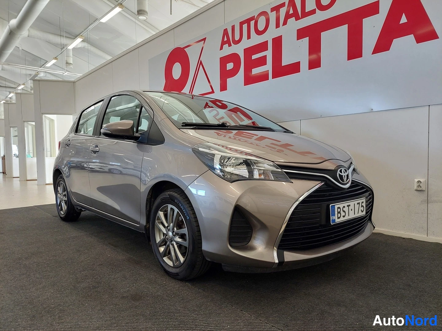 Etuviisto, nokka oikealle: Toyota Yaris -henkilöauton etunurkkakuva sisätiloissa; etusäleikkö, Toyota-merkki ja rekisterikilpi näkyvissä.