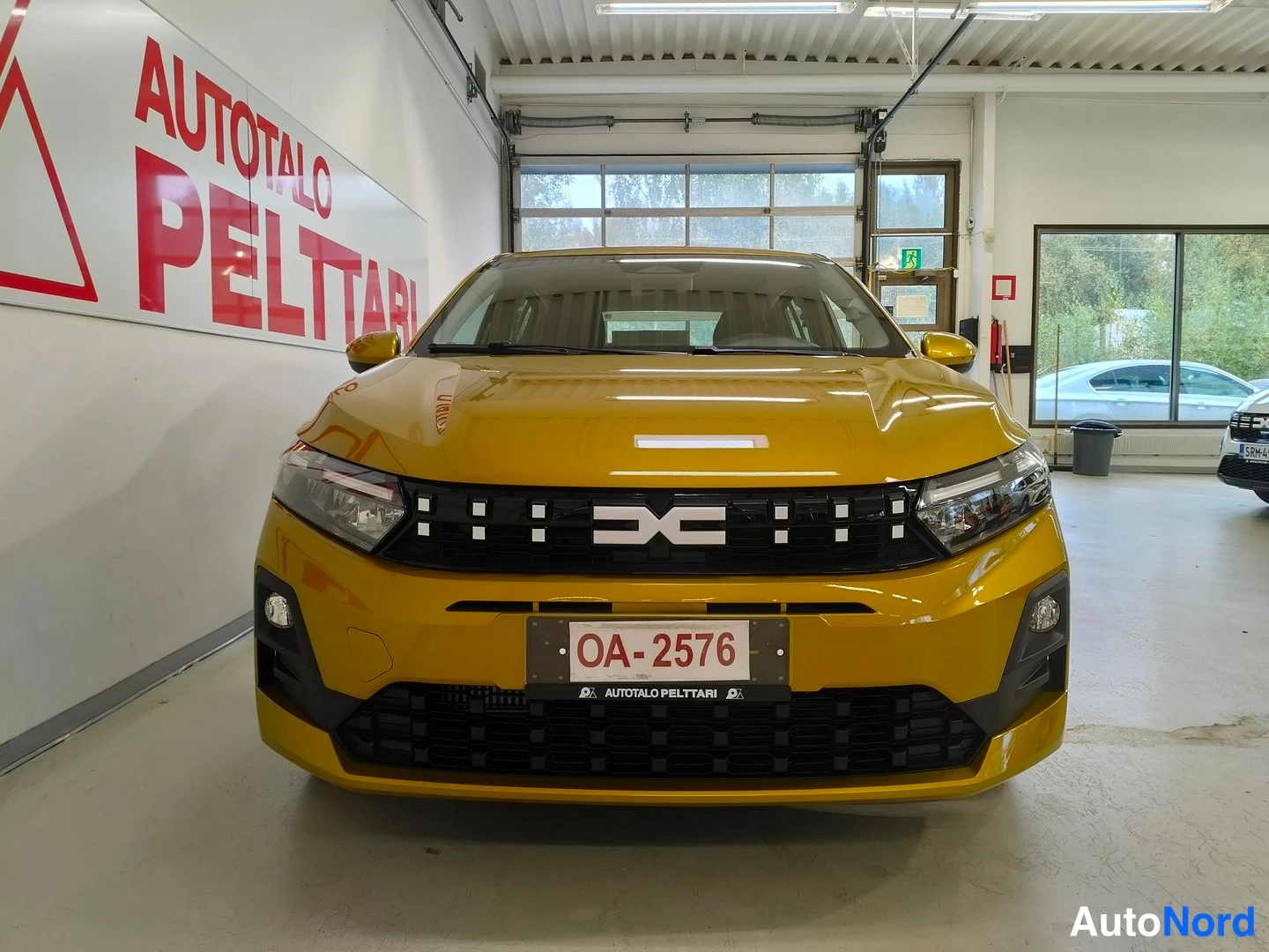 Edestä: Auton etupää frontaalikuvassa sisätiloissa; keltainen kori, Dacia-merkki etusäleikössä ja rekisterikilpi OA-2576.