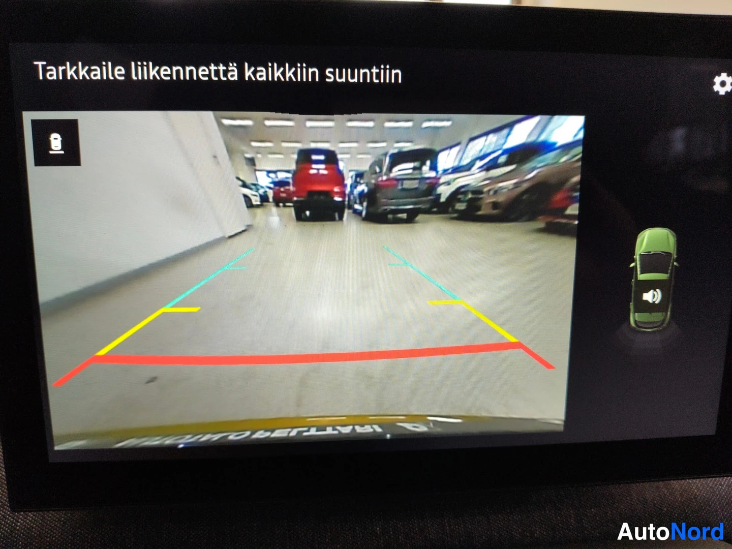Muu: Keskitorvesta kamera otettu kuva näytöllä varastohallissa, tekstinä 'Tarkkaile liikennettä kaikkiin suuntiin' ja apuviivoin.