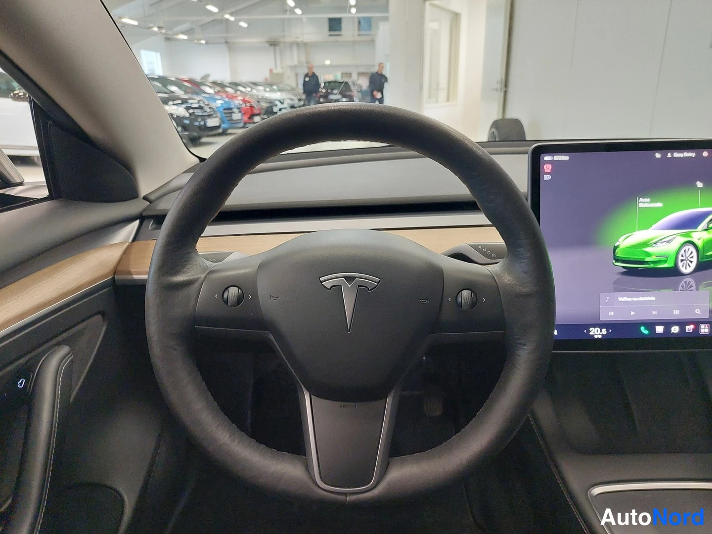 Muu: Auton ohjaamon näkymä: musta Tesla-ohjauspyörä keskellä, puun sävyinen koristepaneeli kojelaudassa ja oikealla kosketusnäyttö, jonka ruudulla näkyy vihreän auto