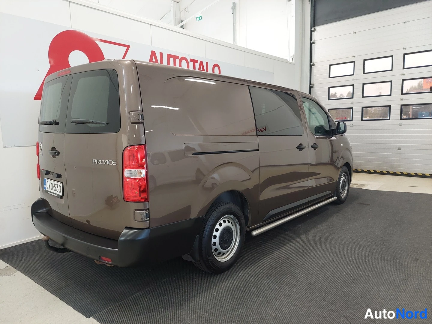Takaviisto: Ruskea Toyota Proace -pakettiauto takaosan ja sivun suunnasta kuvattuna sisätiloissa, PROACE-teksti ja rekisterikilpi näkyvät takaluukussa.