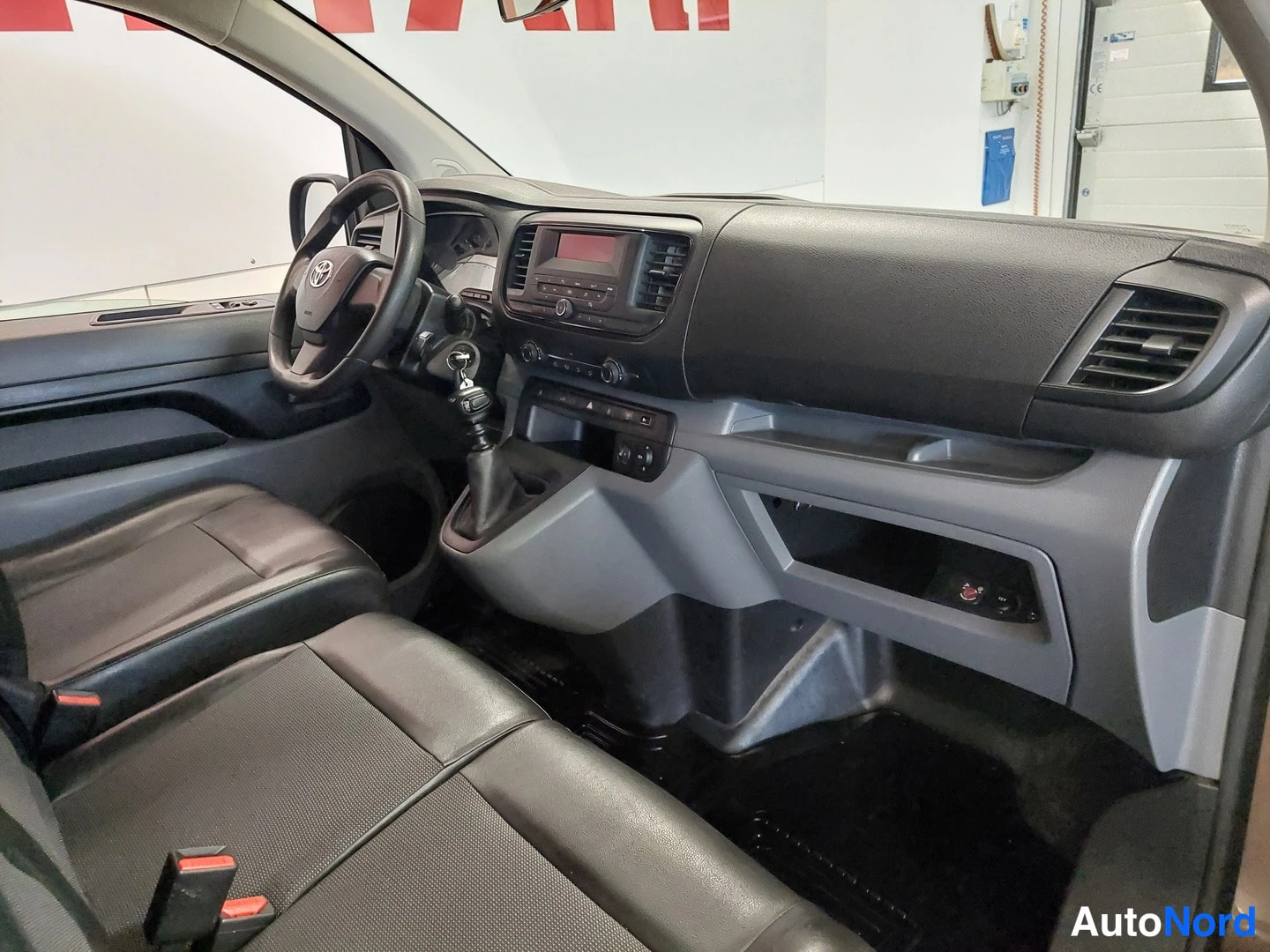 Muu: Auton etuosa sisältä: tumma kojelauta, Toyota-ohjauspyörä, radio paneeli ja manuaalivaihtoehdan vaihteenkeppi.