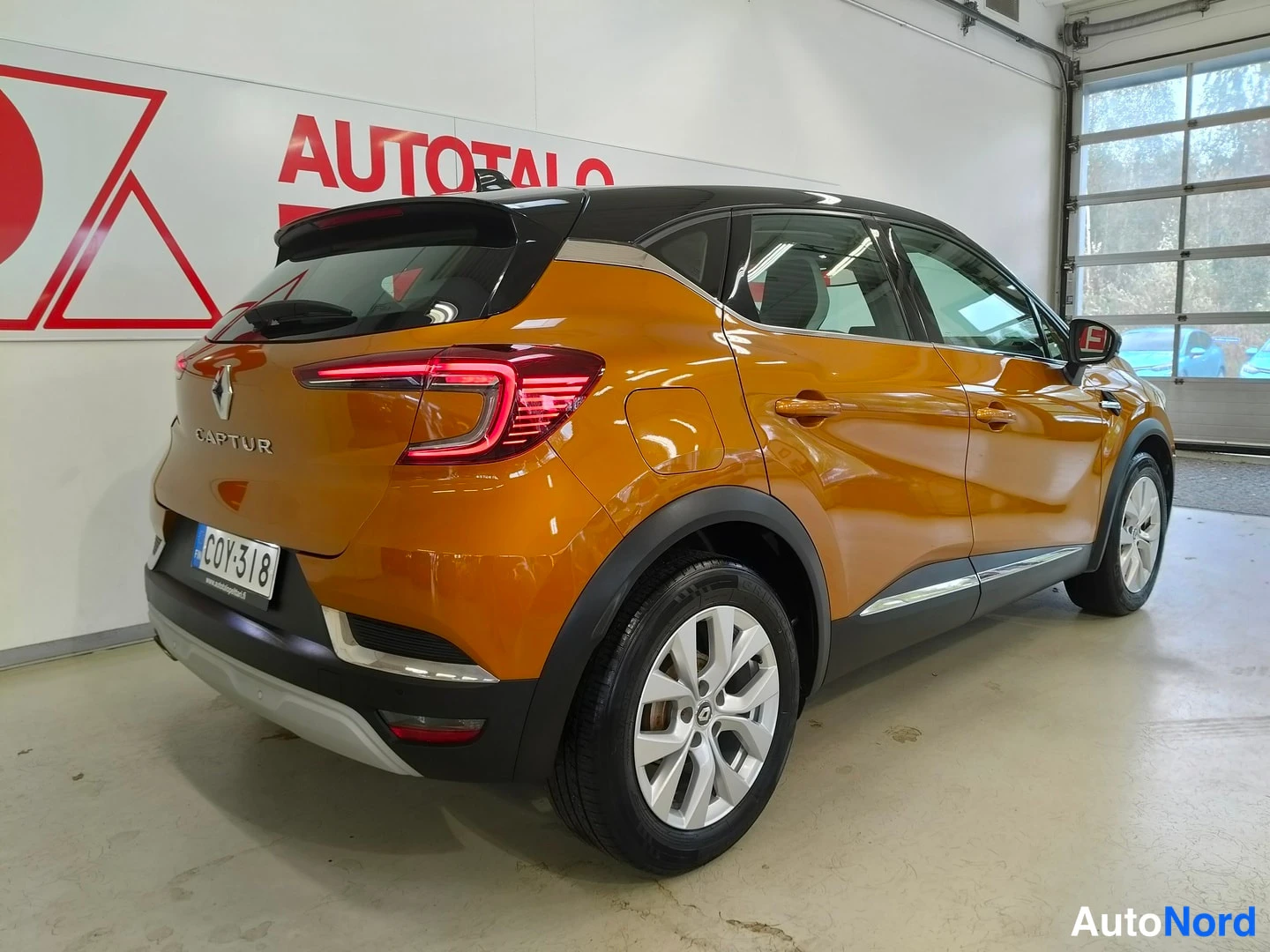 Takaviisto: Oranssi Renault Captur takaa sivusuunnasta kuvattuna sisätiloissa; rekisterikilpi ja CAPTUR-teksti takaluukussa näkyvät.